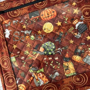 Puede incluir: Un estuche de tela acolchada con una ventana de vinilo transparente que presenta un estampado de Halloween con calabazas, brujas, gatos negros, fantasmas y estrellas sobre un fondo marrón. La palabra Spooky está impresa con una cremallera negra.