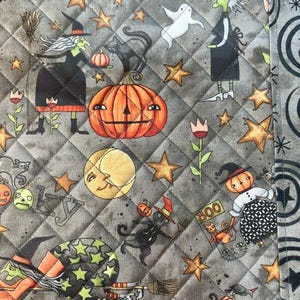 May include: A quilted fabric panel featuring a Halloween pattern with witches, black cats, pumpkins, a ghost, and stars on a gray background. The border displays a repeating circular and star motif. The text BOO is visible on a small sign.