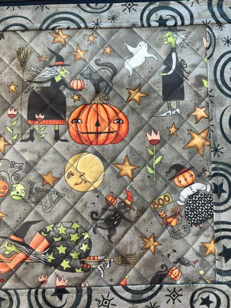 May include: A quilted fabric panel featuring a Halloween pattern with witches, black cats, pumpkins, a ghost, and stars on a gray background. The border displays a repeating circular and star motif. The text BOO is visible on a small sign.