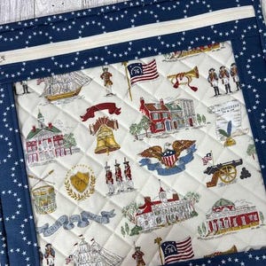May include: A quilted fabric item with a navy blue star-patterned border and a cream-colored center. The center features a patriotic print with historical American imagery, including flags, buildings, and soldiers. A white zipper is visible at the top.