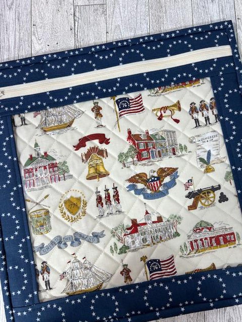 May include: A quilted fabric item with a navy blue star-patterned border and a cream-colored center. The center features a patriotic print with historical American imagery, including flags, buildings, and soldiers. A white zipper is visible at the top.