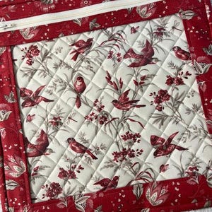 Puede incluir: Una bolsa de tela acolchada rectangular con un borde floral rojo y una cremallera crema. La tela interior presenta un fondo crema con un estampado de pájaros, flores y follaje rojos. La bolsa parece hecha a mano.