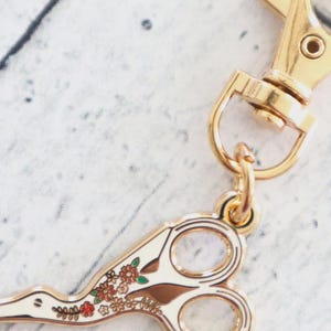 Può includere: Ciondolo a forma di forbice color oro con un design floreale. Il ciondolo è attaccato a un fermaglio color oro. Le forbici hanno uno sfondo bianco con accenti floreali marroni e rossi. Il ciondolo è probabilmente per un portachiavi o un gioiello.