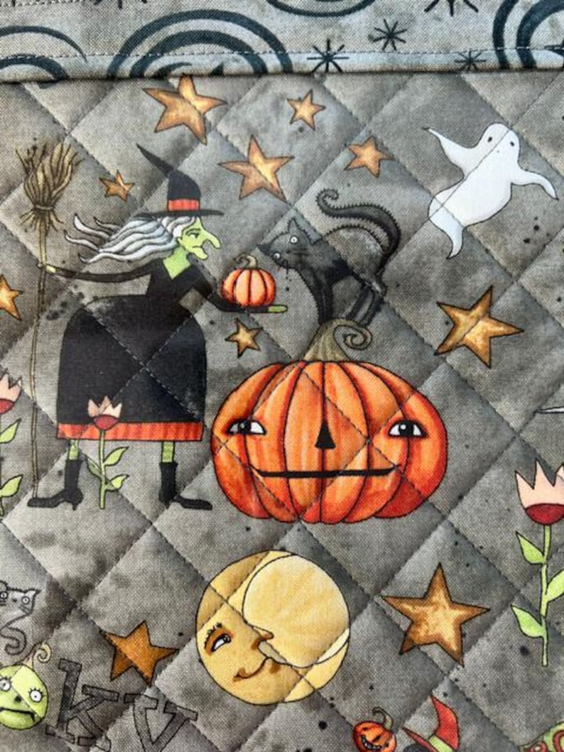 May include: A quilted fabric featuring a Halloween pattern on a gray background. The print includes a green-skinned witch holding a broom and pumpkin, a black cat, a large orange jack-o'-lantern, a white ghost, stars, and a crescent moon.