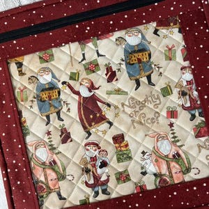 May include: A square quilted fabric pouch with a dark red border featuring white speckles and a black zipper. The centre panel displays a cream-coloured fabric with a repeating pattern of Santa Claus figures, gift boxes, and the text Naughty or Nice?