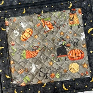 May include: A quilted zipped pouch featuring a black border with yellow crescent moons and stars. The centre panel displays a grey background with a Halloween pattern of witches, pumpkins, ghosts, black cats, and the word Spooky in script.