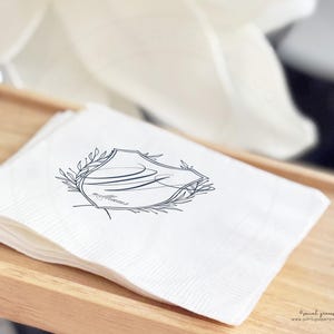 May include: A stack of white paper napkins with a navy blue crest design. The crest features a stylized initial and the word "williams" in script, surrounded by a laurel wreath. The napkins are on a wooden tray.