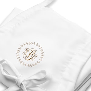 May include: White apron with a gold embroidered monogram inside a laurel wreath. The apron is tied with a white ribbon. The monogram features the letters "EB".