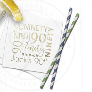 May include: White square paper cocktail napkin with gold text that reads "90 Ninety" and "Jack's 90th". Two striped paper straws, one blue and white, the other green and white, are next to the napkin. A lemon slice is in the upper left corner.