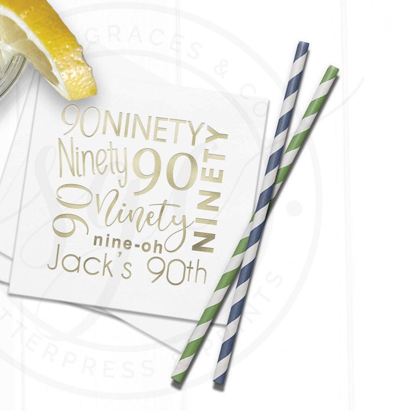 May include: White square paper cocktail napkin with gold text that reads "90 Ninety" and "Jack's 90th". Two striped paper straws, one blue and white, the other green and white, are next to the napkin. A lemon slice is in the upper left corner.