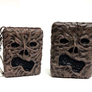 Puede incluir: Dos llaveros rectangulares de textura marrón con un diseño de cara de miedo. El llavero más pequeño tiene una cadena plateada.