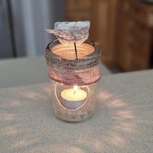 May include: A glass jar candle holder with a burlap wrap and a seashell on top. The candle is lit and the jar is filled with sand.