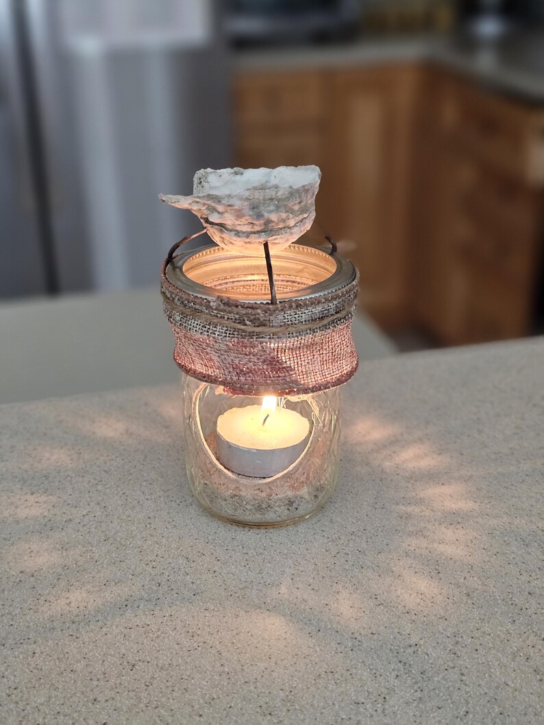May include: A glass jar candle holder with a burlap wrap and a seashell on top. The candle is lit and the jar is filled with sand.