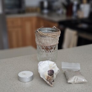 May include: A clear glass jar with a burlap ribbon around it, a white oyster shell, and a small white candle. There is also a small bag of sand.