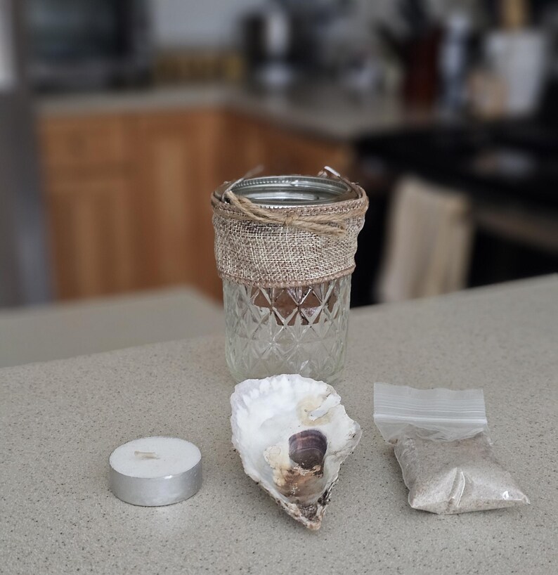 May include: A clear glass jar with a burlap ribbon around it, a white oyster shell, and a small white candle. There is also a small bag of sand.