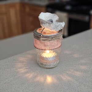 May include: A glass jar candle holder decorated with burlap and a seashell. The candle is lit and the light shines through the jar.