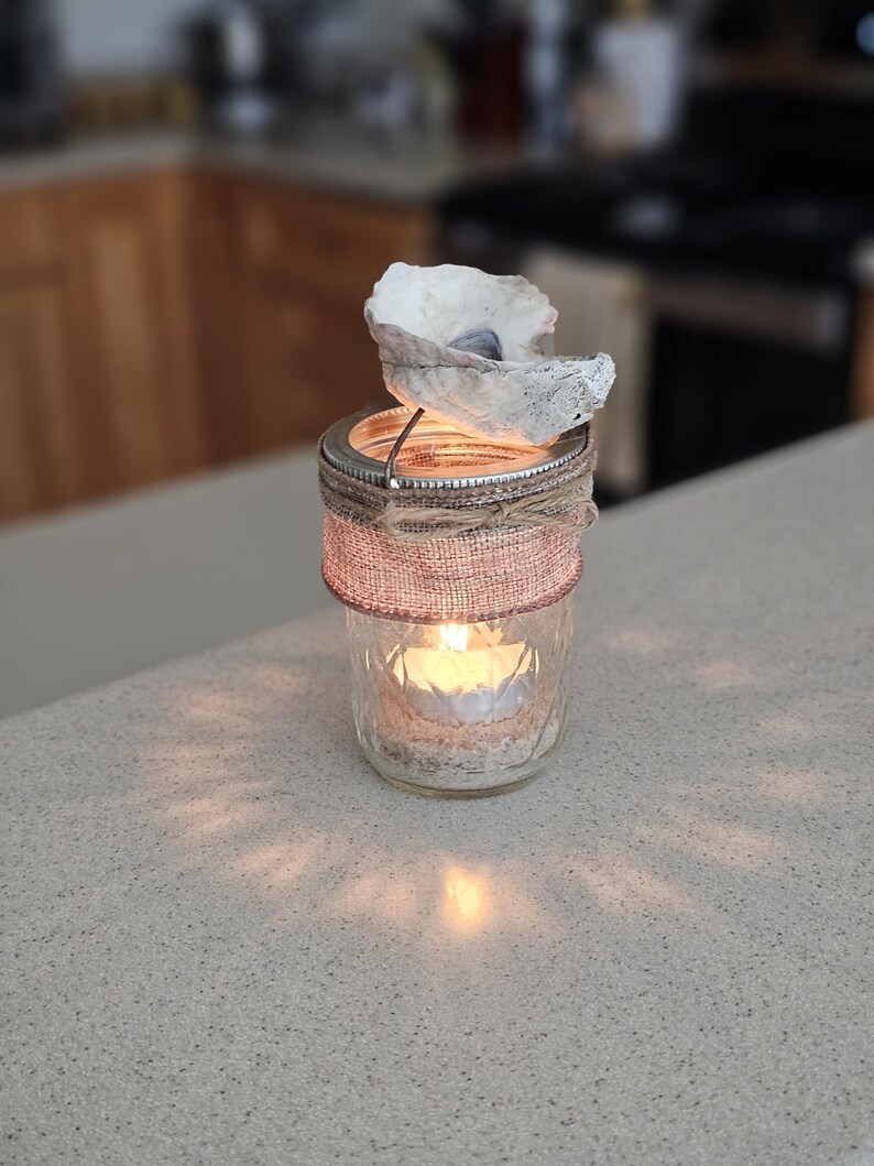 May include: A glass jar candle holder decorated with burlap and a seashell. The candle is lit and the light shines through the jar.