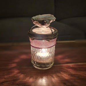 May include: A glass candle holder with a decorative burlap and red ribbon trim. A seashell sits on top of the holder, and a candle is lit inside.