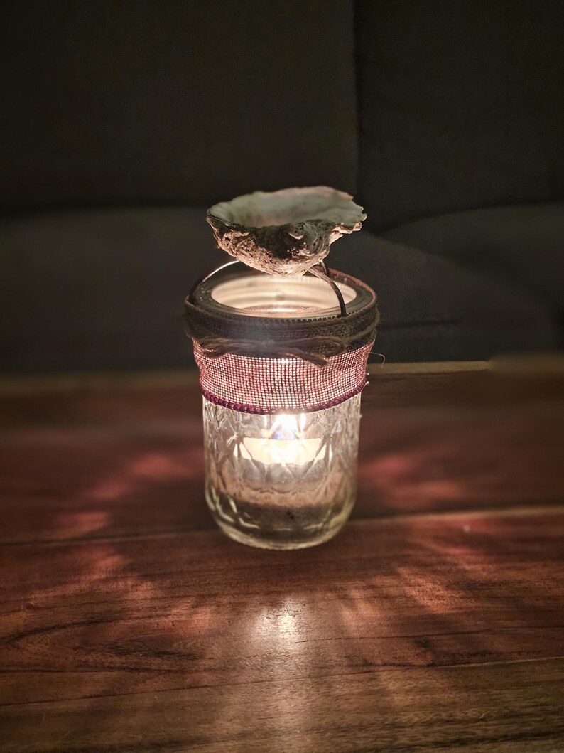 May include: A glass candle holder with a decorative burlap and red ribbon trim. A seashell sits on top of the holder, and a candle is lit inside.