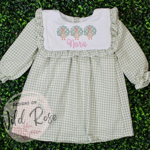 May include: A light green and white gingham dress with long sleeves and a ruffled collar. The white bib features embroidered turkeys and the name "Nora" in pink script. The dress is from "Designs on Wild Rose".