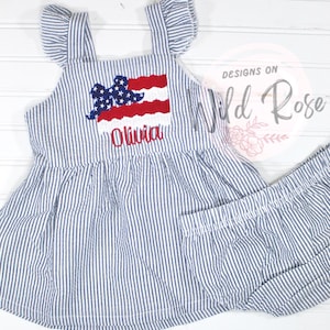Könnte beinhalten: Ein blaues und weiß gestreiftes Seersucker-Kleid mit einer aufgestickten amerikanischen Flagge in Rot, Weiß und Blau und dem Namen "Olivia" auf der Vorderseite. Das Kleid hat Rüschenärmel und wird mit passenden Shorts geliefert.