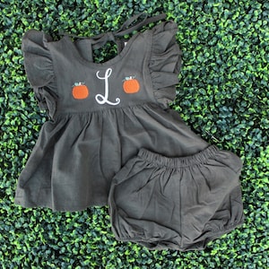 Puede incluir: Un conjunto de bebé niña gris oscuro con una blusa con volantes y bombachos. La blusa tiene calabazas bordadas y la letra "L".