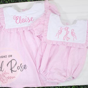 Könnte beinhalten: Zwei rosa Gingham-Kleider mit weißen Kragen und Rüschenbesatz. Das größere Kleid hat den Namen "Eloise" auf dem Kragen gestickt. Das kleinere Kleid hat ein Monogramm auf dem Kragen.