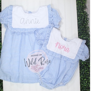 Peut inclure: Deux tenues de bébé en vichy bleu et blanc avec des cols blancs. La plus grande tenue a le nom "Annie" brodé sur le col. La plus petite tenue a le nom "Nora" brodé sur le col. Les tenues sont de Designs on Wild Rose.