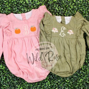 May include: Two baby rompers, one pink and one olive green, with long sleeves and ruffled details. The pink romper has orange pumpkin embroidery and a white monogram. The green romper features floral embroidery and a white monogram.
