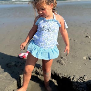 Può includere: Una bambina indossa un costume da bagno floreale blu e bianco con un volant blu a quadri sul fondo. Il costume da bagno ha il nome "Sadie" ricamato sul davanti. La bambina è in piedi su una spiaggia sabbiosa.