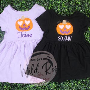 Peut inclure: Deux robes pour enfants, une lavande et une noire, avec un motif de citrouille. La citrouille a des yeux en forme de cœur et une bouche violette. La robe lavande porte le nom "Eloise" brodé, tandis que la robe noire dit "Sadie".
