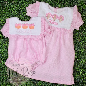 Könnte beinhalten: Zwei passende rosa Gingham-Kleider. Eines ist ein Strampler mit dem Namen "Eleanor" auf dem Lätzchen, das andere ist ein Kleid mit dem Namen "Nora" auf dem Lätzchen. Beide haben Rüschenärmel und Lätzchen mit Kürbis- und Truthahnstickereien.