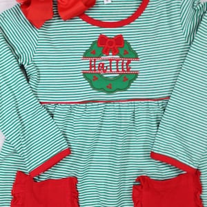 Può includere: Abito a maniche lunghe a righe verdi e bianche con bordi rossi. L'abito ha un fiocco rosso e una ghirlanda verde con il nome "Hallie" ricamato. L'abito ha due tasche rosse con volant.