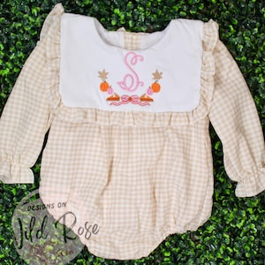 May include: A beige and white gingham baby romper with long sleeves and a white bib collar. The collar features an embroidered pink initial "S" surrounded by pumpkins and fall foliage. The text "DESIGNS ON Wild Rose" is visible.