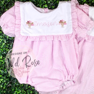 Peut inclure: Barboteuse pour bébé à carreaux roses et blancs avec un bavoir blanc. Le bavoir est brodé du nom "Emerson" et de deux motifs de dindes. La barboteuse a des manches et des bordures à volants. Le texte "Designs on Wild Rose" est visible.