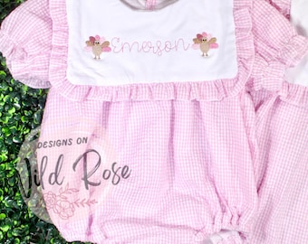 May include: Pink and white gingham baby romper with a white bib. The bib is embroidered with the name "Emerson" and two turkey designs. The romper has ruffled sleeves and trim. The text "Designs on Wild Rose" is visible.