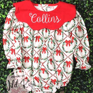 May include: A white and red Christmas-themed baby romper with long sleeves and a Peter Pan collar. The romper features a pattern of wreaths and red bows. The collar is red with the name "Collins" embroidered in white.