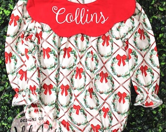 May include: A festive baby romper in white with a pattern of Christmas wreaths and red bows. It has long sleeves and a red Peter Pan collar embroidered with the name "Collins". The romper is designed for a baby.