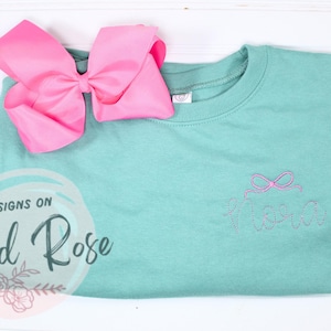 May include: A teal sweatshirt with a pink bow and the word "Missy" embroidered on the front. The sweatshirt is folded and lying on a white surface. A pink bow is also lying on the surface.