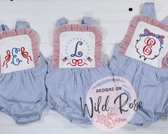 May include: Three blue and white gingham baby rompers with red and white ruffled trim. Each romper has a white embroidered patch with a different letter and patriotic design. The patches feature red, white, and blue stars, flags, and bows.