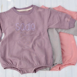 Könnte beinhalten: Drei langärmelige Baby-Bodys in verschiedenen Farben. Der Body oben ist lila mit dem Namen "Sadie" auf der Vorderseite bestickt.