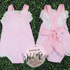 Puede incluir: Unisie de beb&eacute; de gingham rosa con ribete de encaje blanco y letras bordadas. El body tiene un lazo rosa en la espalda y botones blancos.
