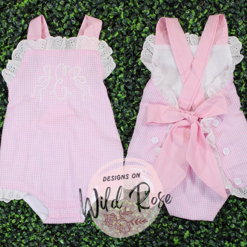 Puede incluir: Unisie de beb&eacute; de gingham rosa con ribete de encaje blanco y letras bordadas. El body tiene un lazo rosa en la espalda y botones blancos.