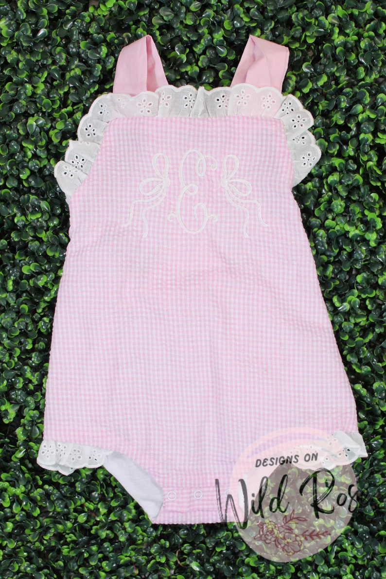 Puede incluir: Un body de beb&eacute; de gingham rosa y blanco con ribete de encaje blanco y un monograma bordado blanco. El body tiene un cuello con volantes y est&aacute; hecho de una tela suave y c&oacute;moda. El body es de Designs on Wild Rose.