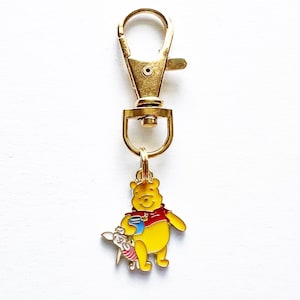 May include: A gold-colored keychain clip with a yellow Winnie the Pooh charm wearing a red shirt and holding a blue pot. A small Piglet charm is also attached. The background is white.