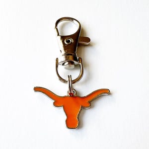 May include: A keychain featuring a burnt orange longhorn symbol with a silver-colored metal clasp. The longhorn is a stylized representation of a bull's horns, with a rounded body and curved horns. The keychain is designed to attach to keys or bags.