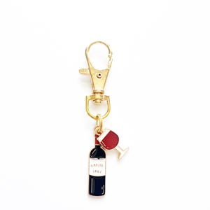 May include: Gold keychain with a red wine bottle and glass charm. The bottle has a label that reads "Lafite 1982".