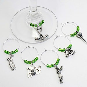 May include: Six green and silver wine glass charms with golf themed charms. The charms include a golf club, a golf cart, a golfer, a golf bag, a golf ball, and a golf tee.