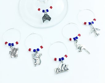 May include: Set of six Australian themed wine glass charms. Each charm features a different Australian icon, including a koala, kangaroo, Sydney Opera House, and the word "Australia". The charms are made of metal and have red, white, and blue beads.