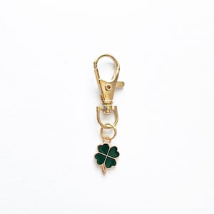 May include: A gold-colored metal clip with a small, four-leaf clover charm. The clover is dark green with gold-colored trim. The clip is designed to attach to a keychain or other items.